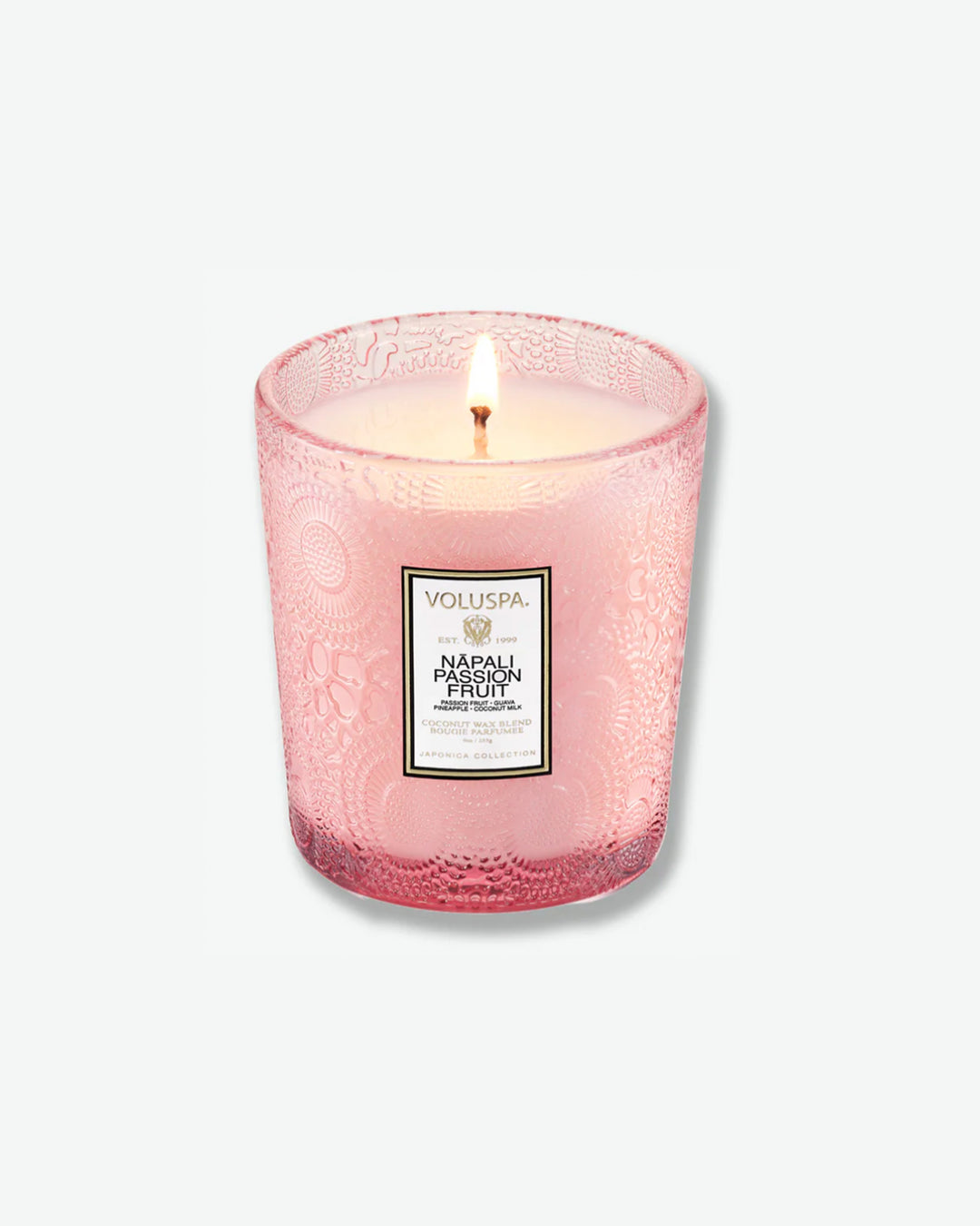 High-angle product shot of a burning Voluspa Nāpali Passion Fruit classic textured glass candle against a plain gray background. The single-wick flame burns in the center of the soft pink, embossed glass holder.