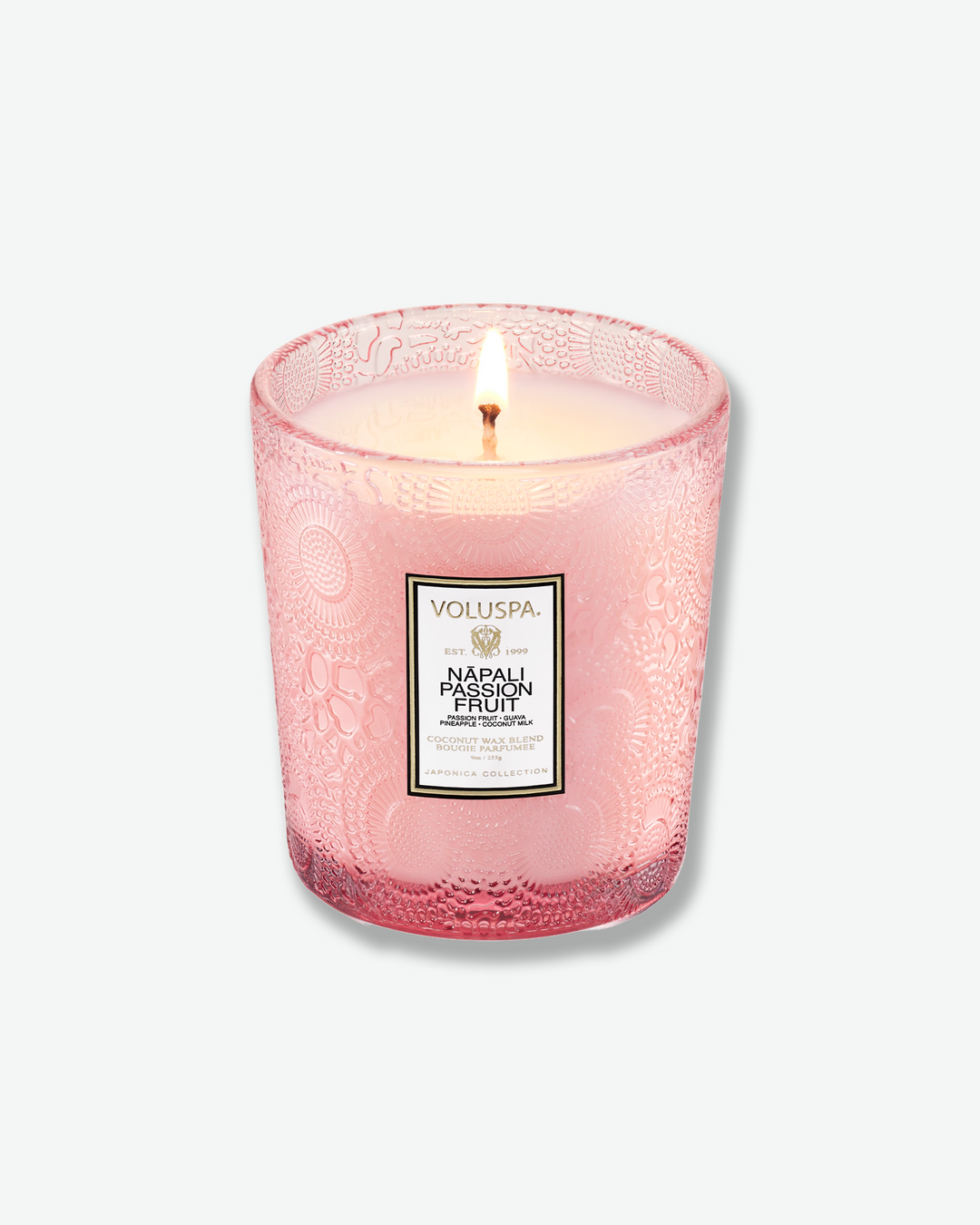 High-angle product shot of a burning Voluspa Nāpali Passion Fruit classic textured glass candle against a plain gray background. The single-wick flame burns in the center of the soft pink, embossed glass holder.