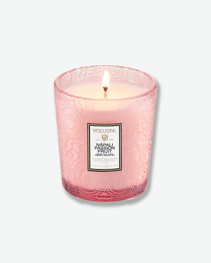 High-angle product shot of a burning Voluspa Nāpali Passion Fruit classic textured glass candle against a plain gray background. The single-wick flame burns in the center of the soft pink, embossed glass holder.