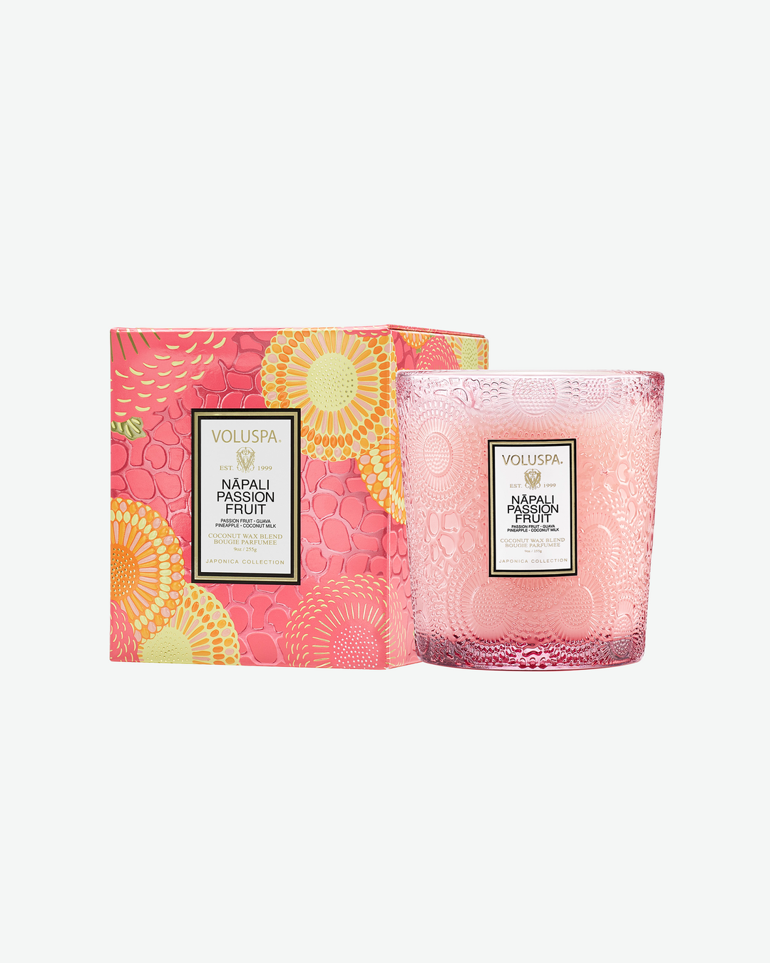 Product shot of a Voluspa Nāpali Passion Fruit classic textured glass candle standing next to its matching square outer packaging. The light pink tapered glass jar sits beside a box featuring a vibrant pink, orange, and yellow botanical floral design.