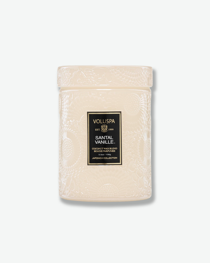 Front view of a Voluspa Santal Vanille small jar candle against a plain gray background. The candle features a cream-tinted glass body completely embossed with swirling floral designs, finished with a black and gold central label and a matching textured glass screw-on lid.