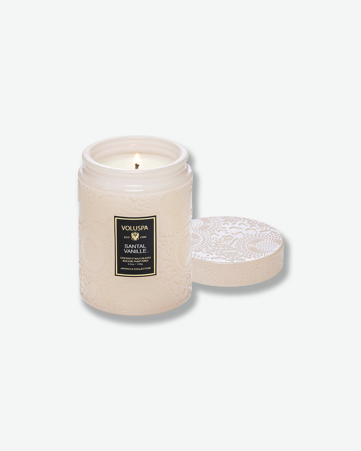 Product shot of a lit Voluspa Santal Vanille small glass jar candle. The single wick is burning inside the cream-tinted embossed container, with its matching textured glass lid resting face-up next to the jar.