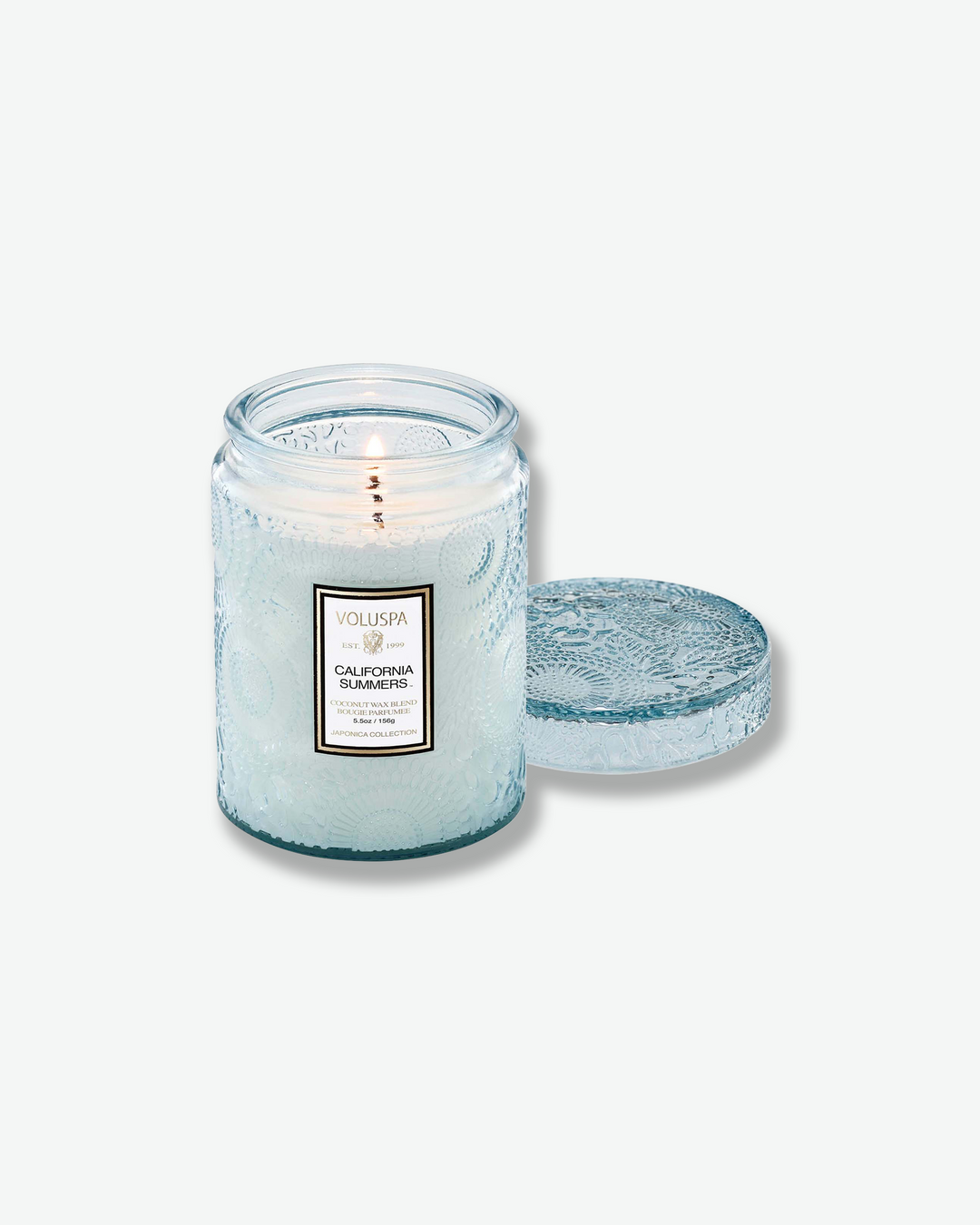 Product shot of a lit Voluspa California Summers small glass jar candle. A single-wick flame glows within the pale blue embossed jar, and its matching textured glass lid rests flat on the surface to the right.