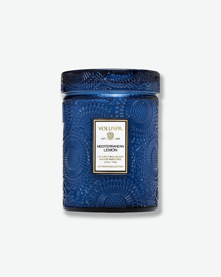 Front view of a Voluspa Mediterranean Lemon small jar candle against a plain gray background. The deep cobalt blue glass jar is intricately embossed with swirling floral textures and capped with a matching textured glass screw-on lid.
