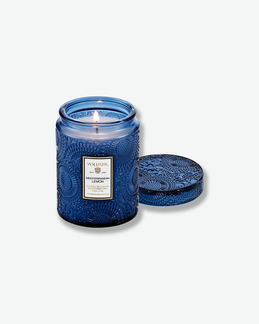 Product shot of a lit Voluspa Mediterranean Lemon small glass jar candle. The flame burns gently inside the cobalt blue embossed glass vessel, with its matching textured blue glass lid resting flat on the surface next to it.
