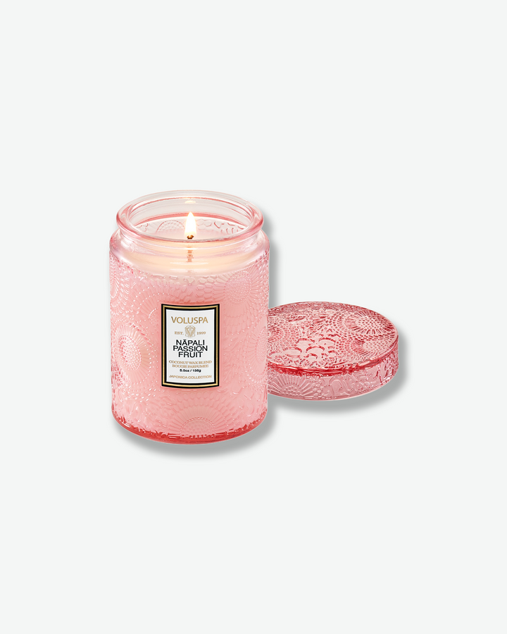 Product shot of a lit Voluspa Nāpali Passion Fruit small glass jar candle. The single wick is burning smoothly inside the soft pink embossed container, with its matching textured pink glass lid resting face-up next to the jar.