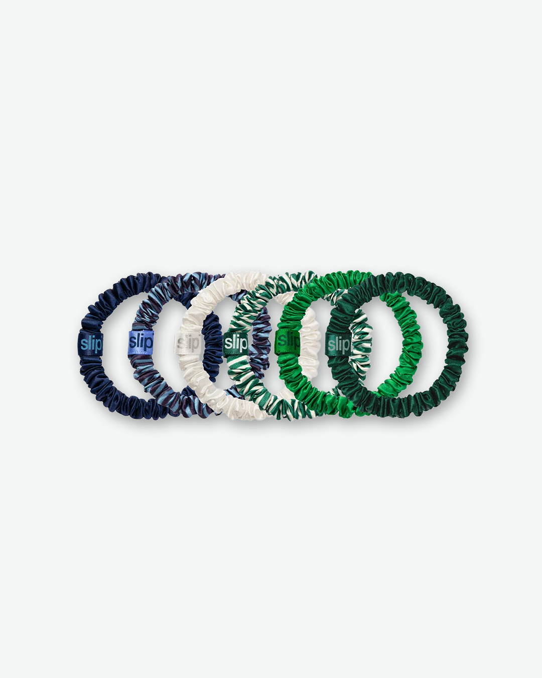 A horizontal lineup of six Slip brand skinny silk scrunchies overlapping each other against a light gray background. From left to right, the colors and patterns are: solid navy blue, a blue and navy abstract patterned print, solid cream, a green and white abstract zebra-like pattern, a vibrant kelly green, and a deep emerald green. Each scrunchie features a small silk fabric tag matching its base color with the word "slip" printed on it.