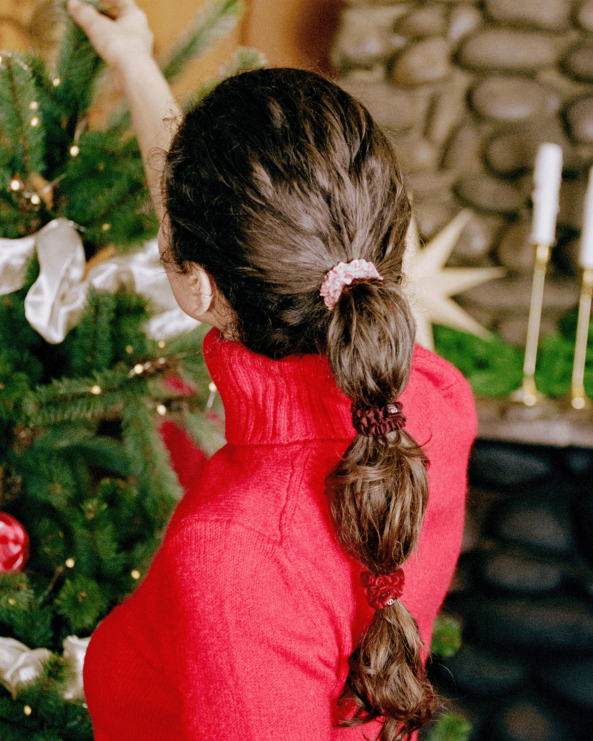 Woman in a red sweater decorating a Christmas tree, with brown hair styled in a bubble ponytail secured with Slip silk skinny scrunchies in pink, burgundy, and red.
