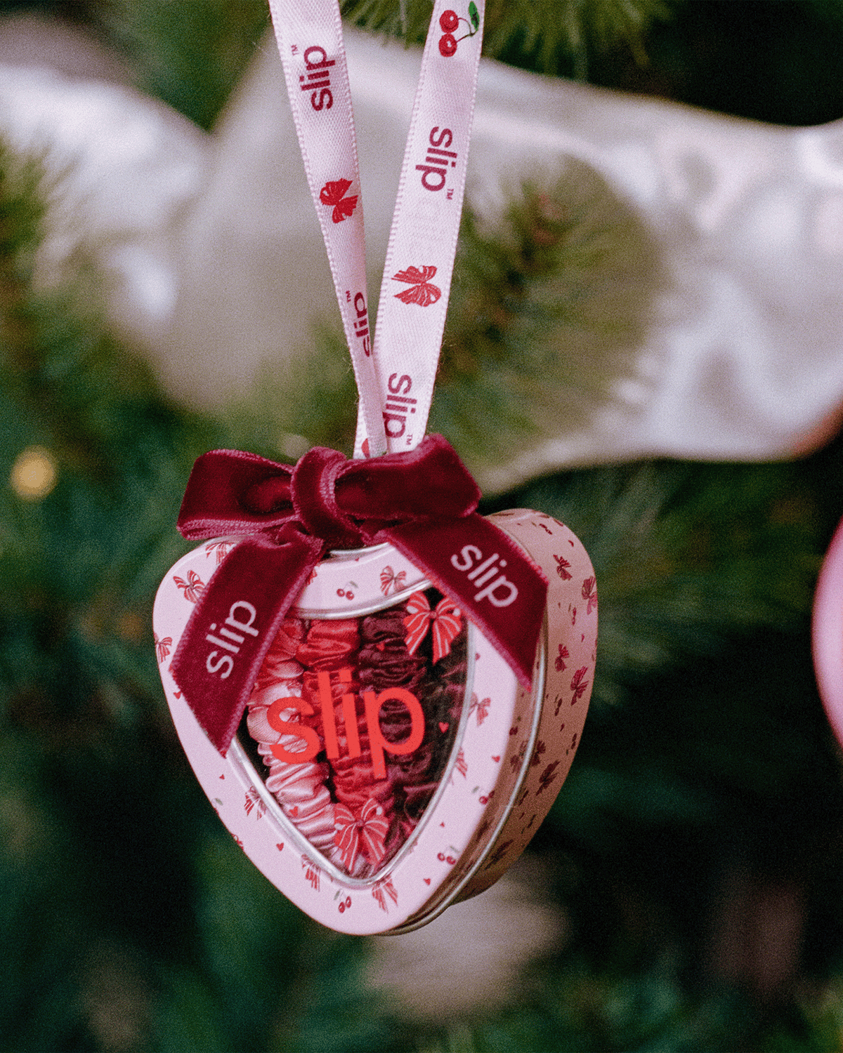 Slip holiday heart-shaped ornament filled with pink, red, and burgundy silk skinny scrunchies, featuring festive cherry and bow illustrations and a burgundy velvet ribbon hanging on a Christmas tree.