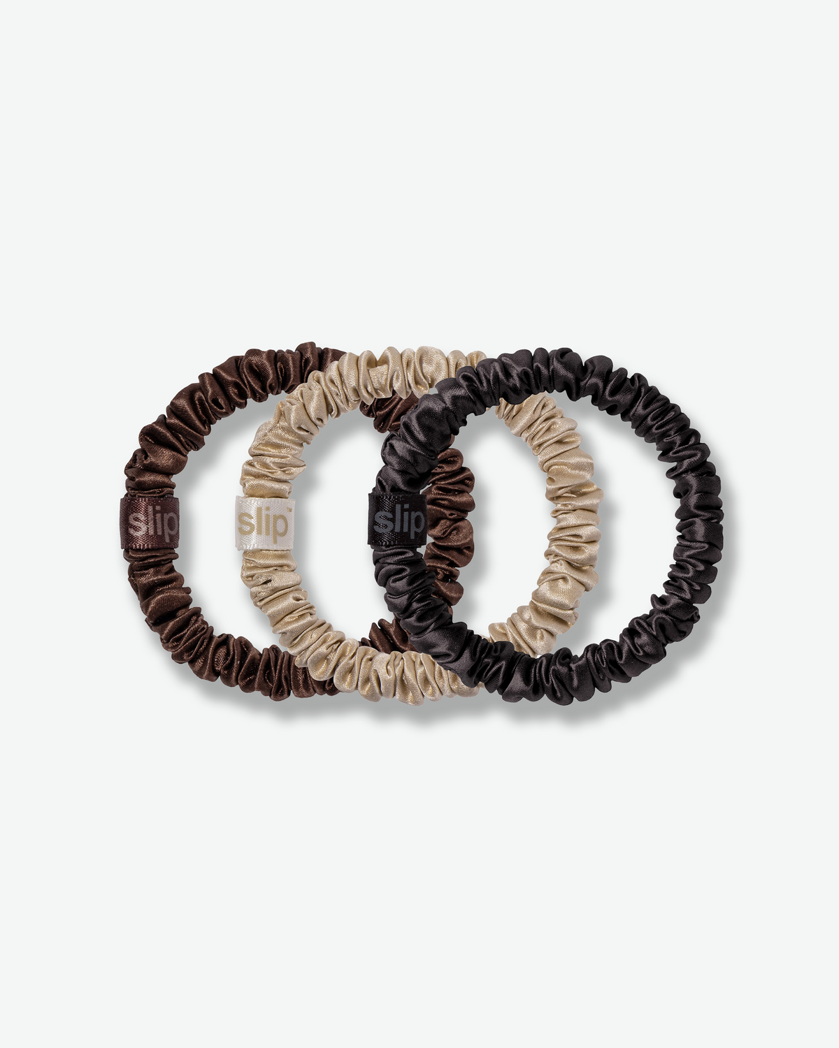 Slip silk skinny scrunchies in chocolate brown, champagne, and black, featuring a smooth silk finish and branded tags on a light gray background.
