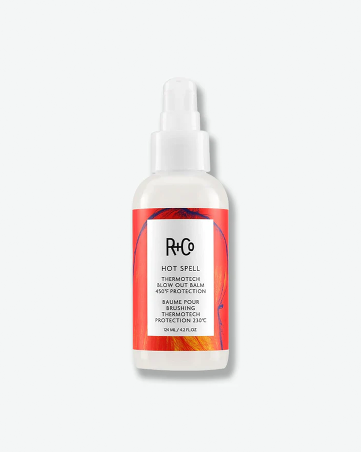 A product shot of a R+Co Hot Spell Thermotech Blow Out Balm bottle, centered against a plain light gray background. The curved bottle is opaque white and is fitted with a matching white pump dispenser and a clear protective cap. The bottle features a vibrant red orange graphic label with a central white square that displays the "R+Co" logo, the product name, and description details including "450°F PROTECTION" and the French translation, ending with the volume "124 ML / 4.2 FL.OZ".