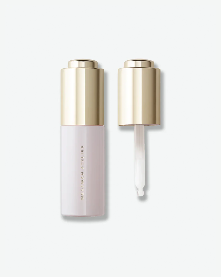 Westman Atelier liquid product in a minimalist pale pink bottle with a metallic gold cap, shown alongside the unscrewed dropper top and a clear glass pipette containing a milky white formula, isolated on a light grey background.