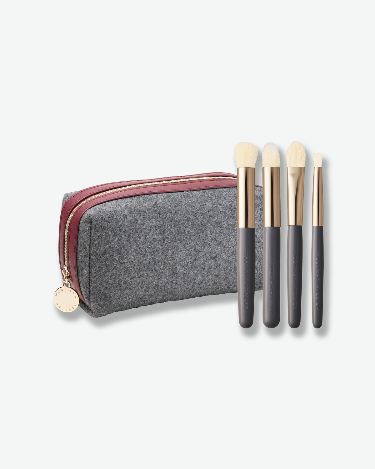Westman Atelier four-piece makeup brush set featuring the Baby Blender Brush, Spot Check Brush, and two Eyeshadow Brushes with gray handles, gold ferrules, and a gray felt travel case with burgundy trim.