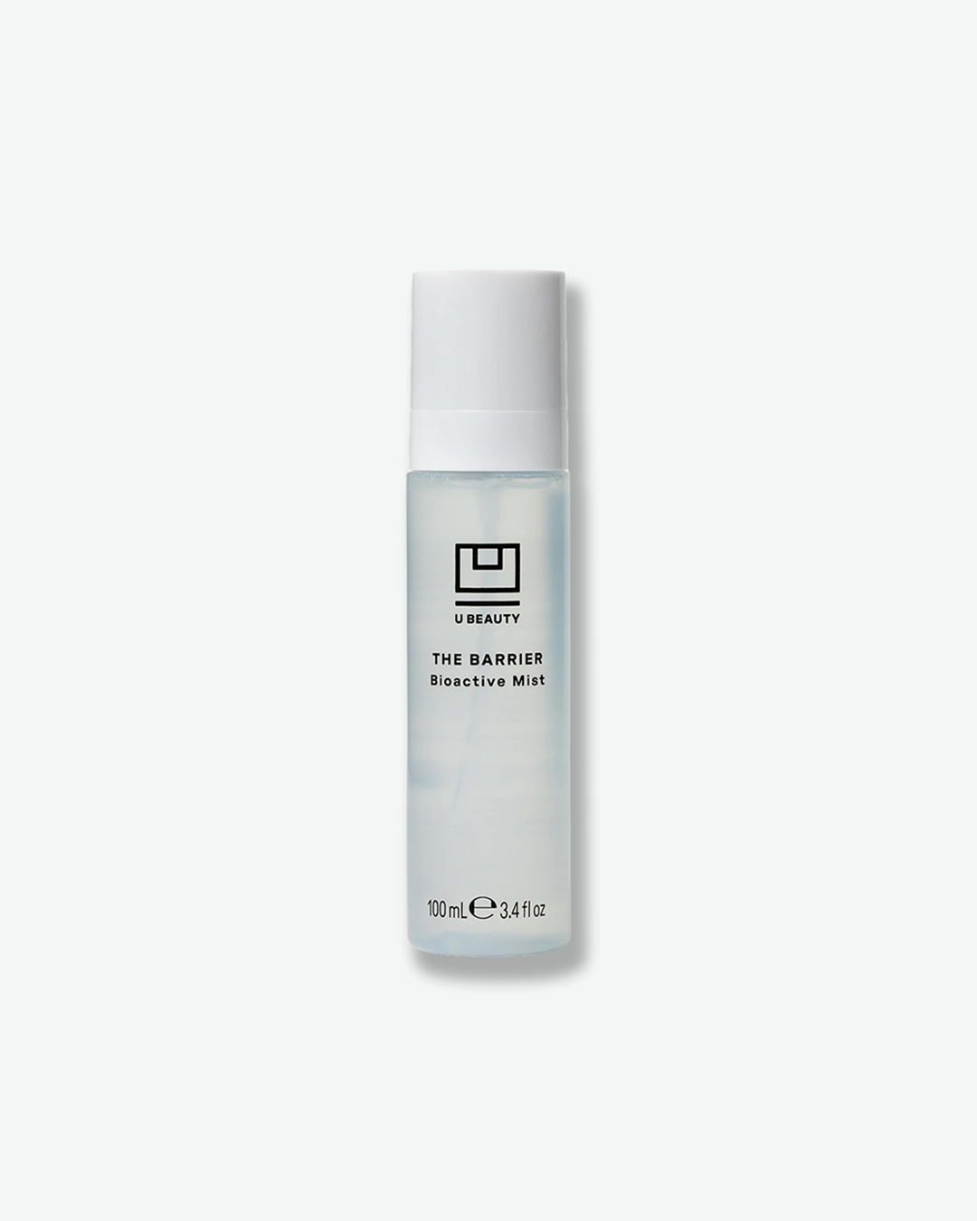 A product shot of a U Beauty The Barrier Bioactive Mist bottle, centered against a plain light gray background. The slender, cylindrical bottle is crafted from frosted clear glass, revealing a subtle, light blue-tinted liquid formula inside, and is fitted with a solid white pump cap. The front features the black logo emblem above the text "THE BARRIER Bioactive Mist", finishing with the volume markings "100mL e 3.4 fl oz" at the bottom.
