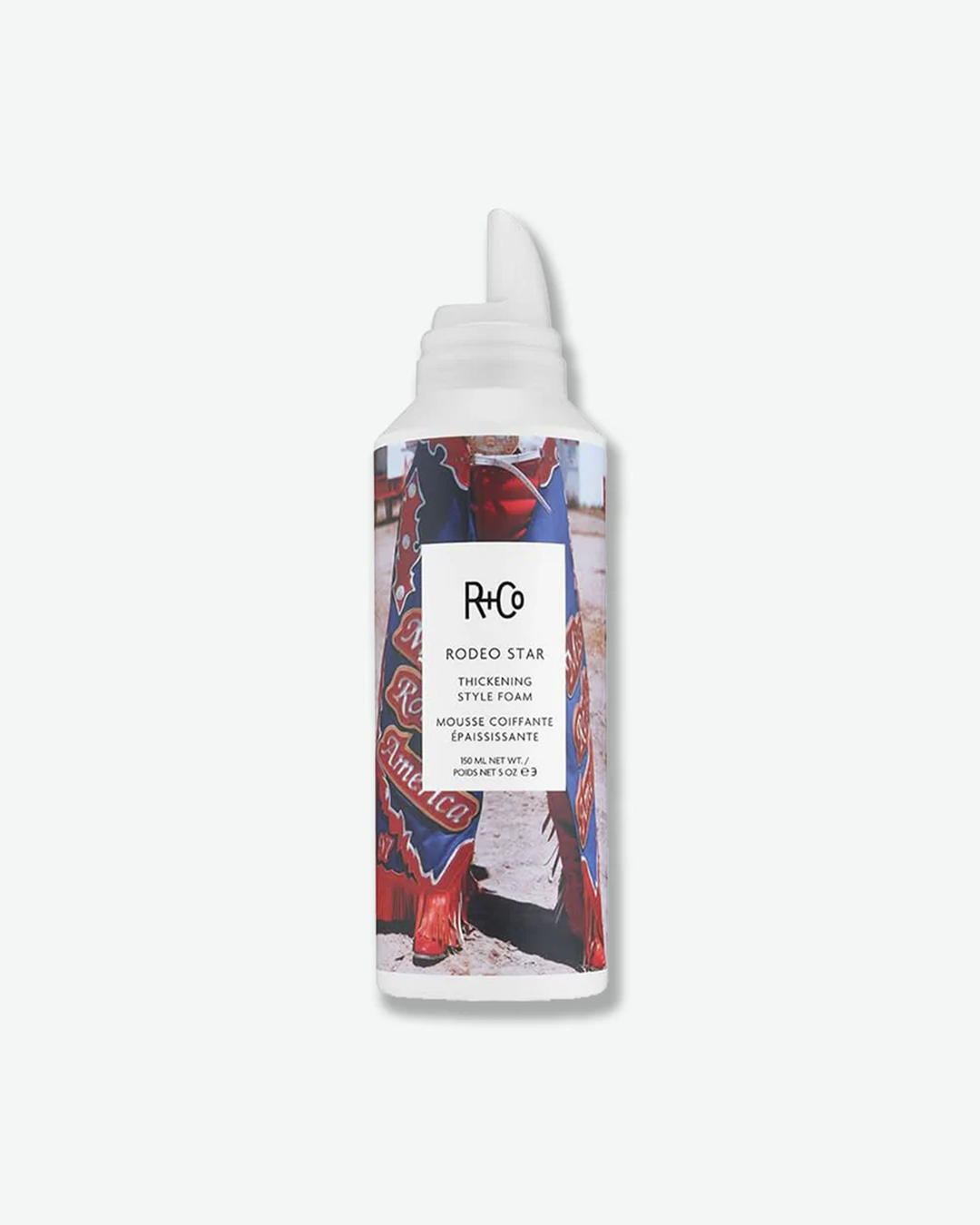 A product shot of a R+Co Rodeo Star Thickening Style Foam canister, centered against a plain light gray background. The cylindrical aluminum canister is wrapped in an photographic label depicting a person wearing ornate cowboy chaps. A central white rectangle overlays the image, featuring the black "R+Co" logo, the product name "RODEO STAR", "THICKENING STYLE FOAM", and its French translation, finished with the volume "150 ML NET WT. / POIDS NET 5 OZ". The bottle is topped with a distinctive white curved fo