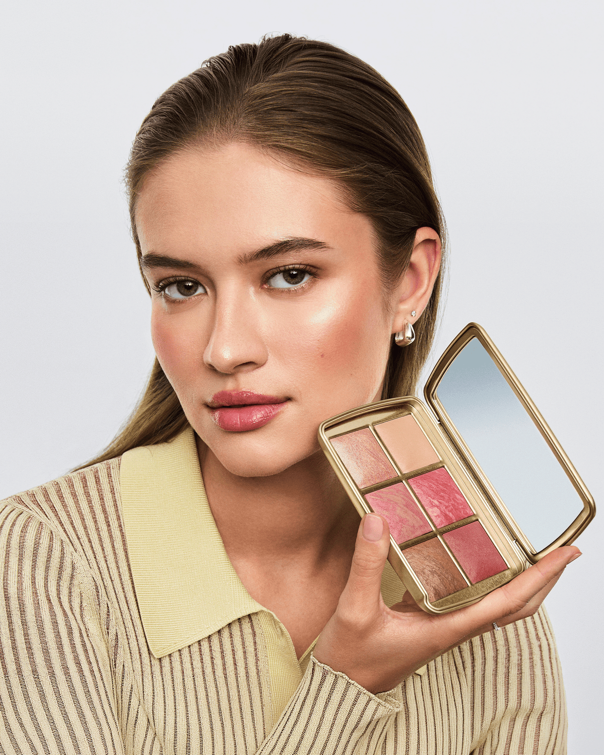Model holding the open Hourglass Ambient Lighting Edit – Unlocked Palette in Hope, showcasing the six complexion shades and gold compact.