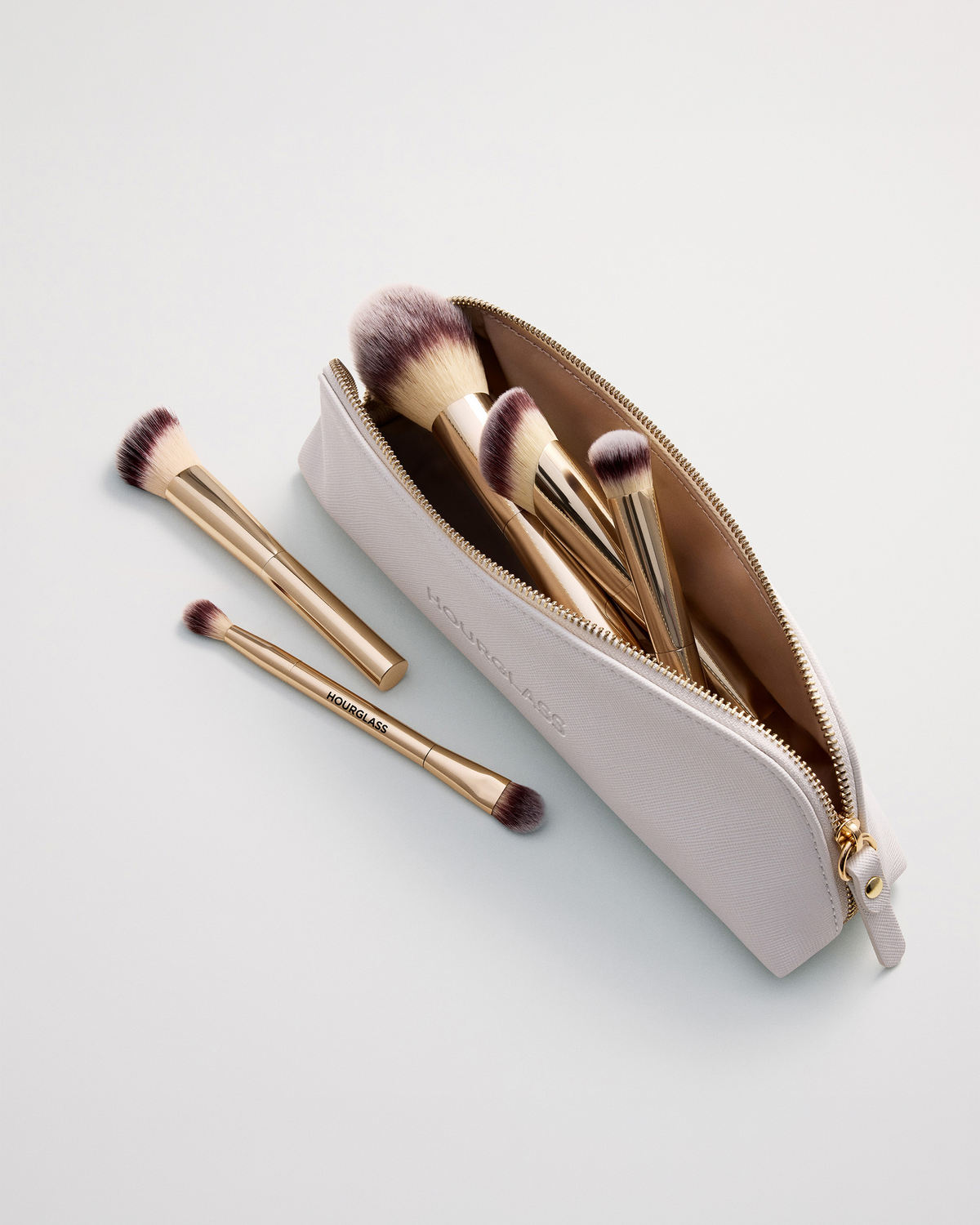 Hourglass travel makeup brushes arranged inside and beside the cream zippered brush case, with soft ivory-to-burgundy bristles and gold handles.