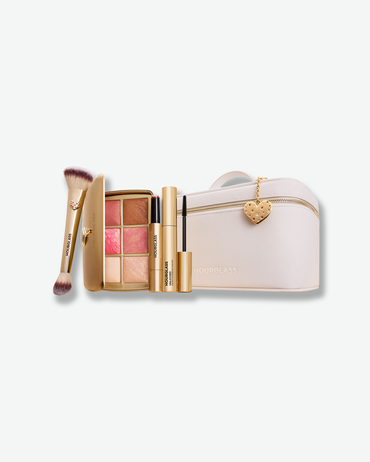Hourglass holiday makeup set featuring a white vanity case, Ambient Lighting Edit palette, dual-ended brush, mascara, and lip product with gold accents and a heart charm.