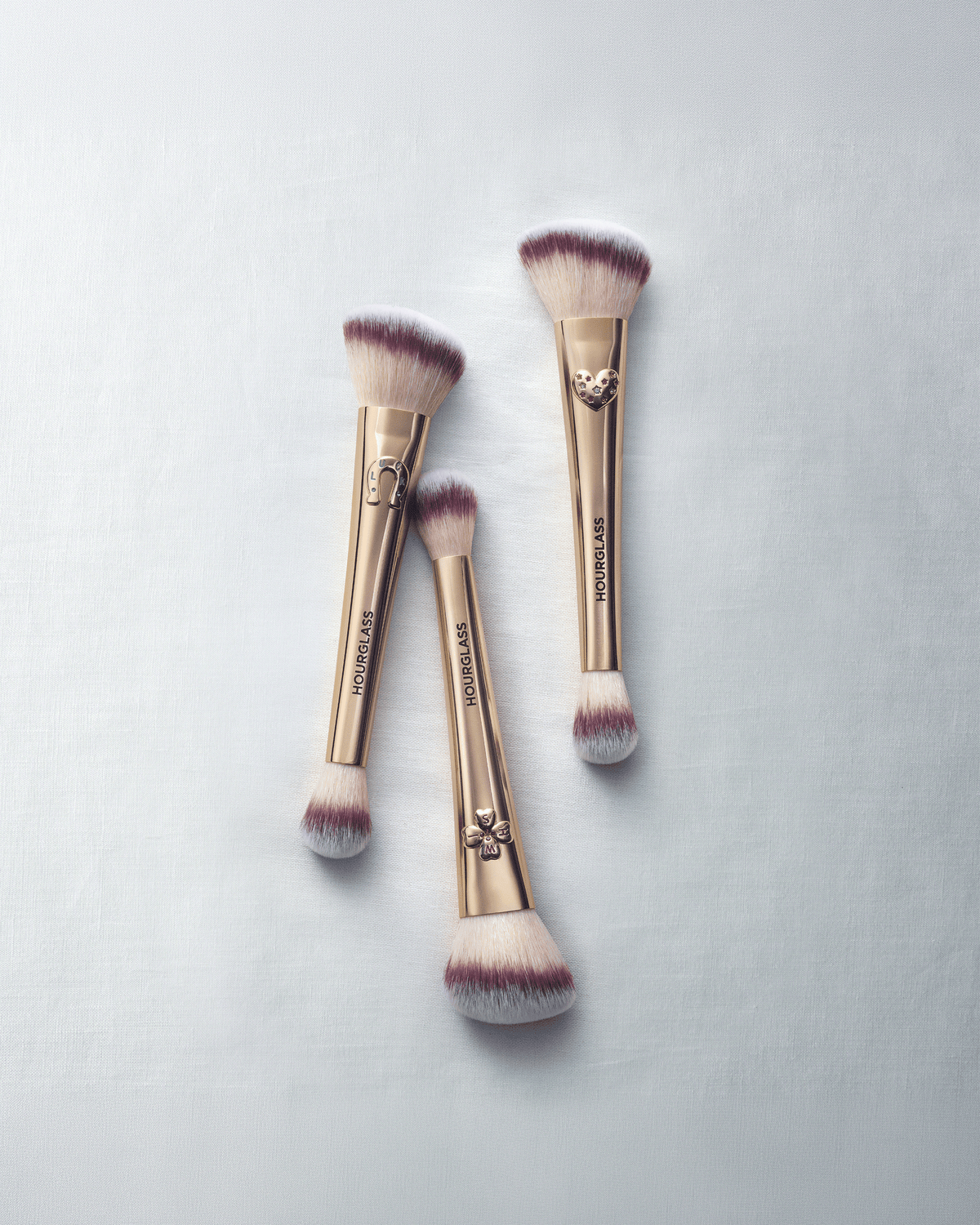 Three limited-edition Hourglass Ambient Lighting Edit Brushes with decorative gold charms arranged on a soft light blue background.