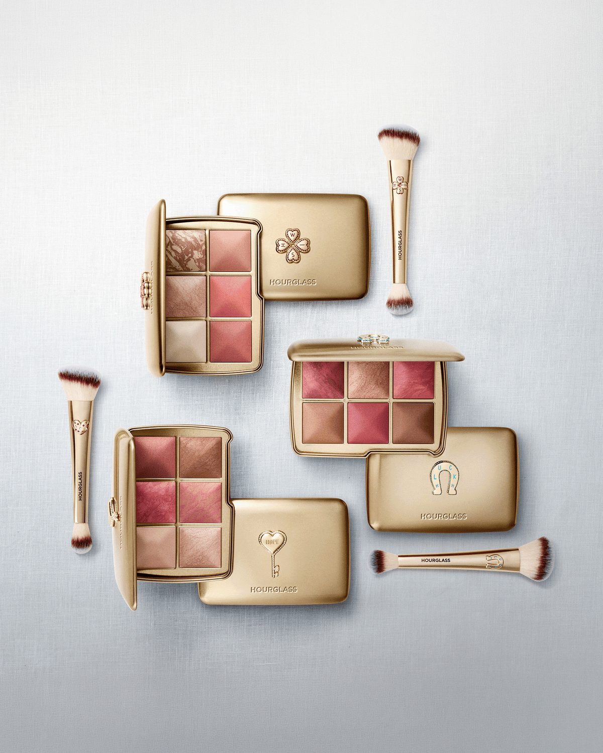 Hourglass holiday makeup collection featuring limited-edition Ambient Lighting palettes and coordinating gold double-ended brushes arranged on a light blue background.