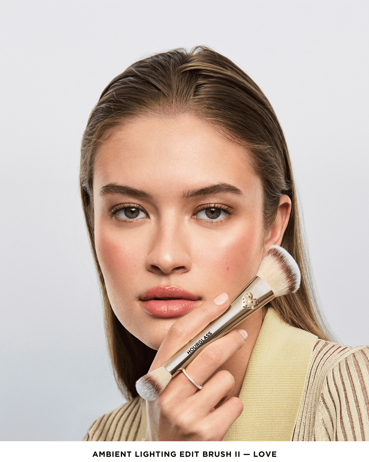 Model holding the Hourglass Ambient Lighting Edit Brush II – Love against her cheek, showcasing the limited-edition gold double-ended design.