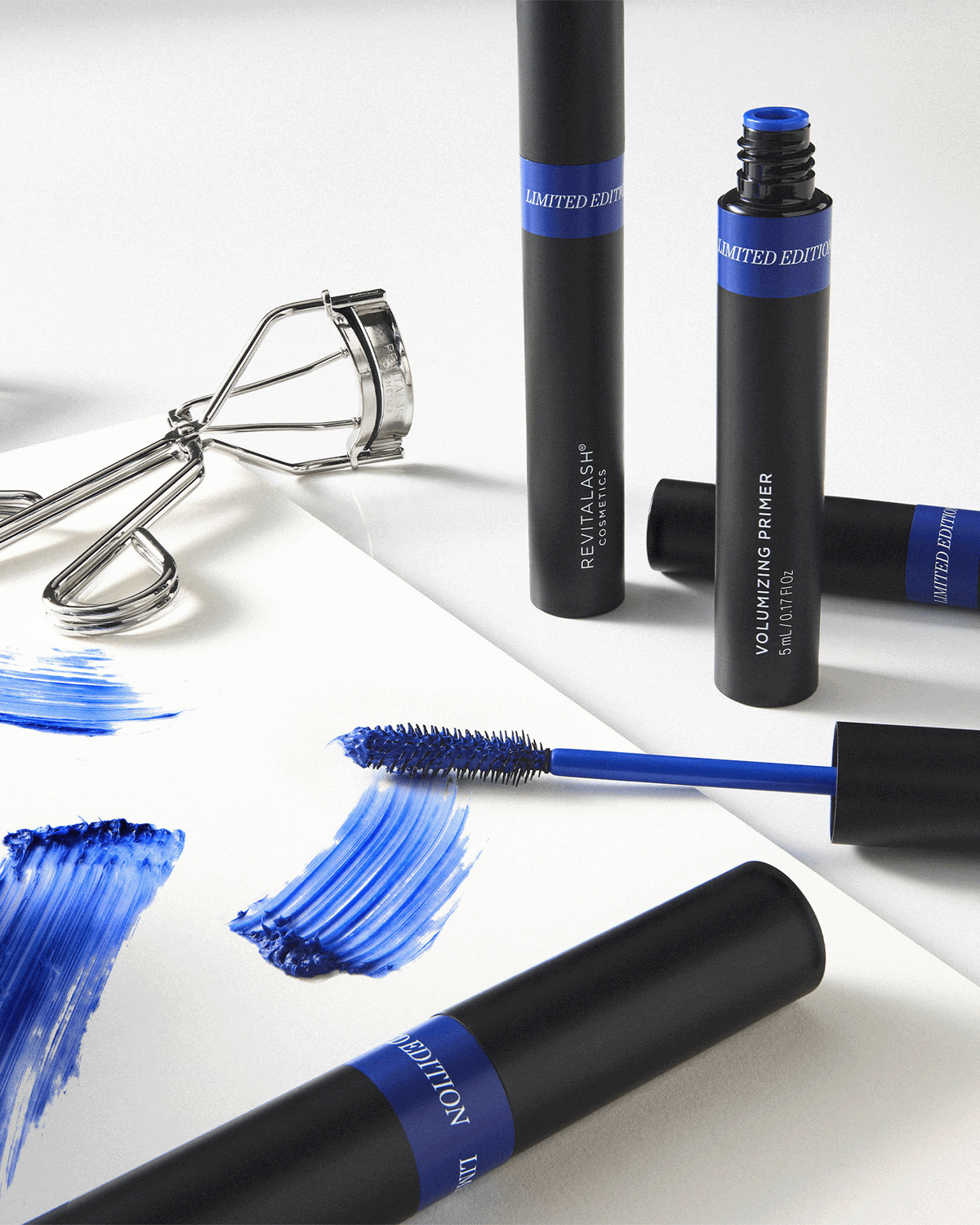 RevitaLash Volumizing Primer, blue applicator wand, and eyelash curler styled with cobalt blue product swatches.