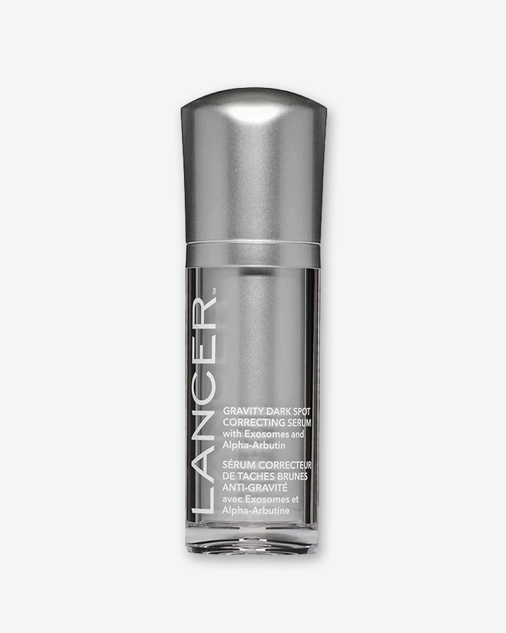 A product shot displaying the front view of a Lancer Gravity Dark Spot Correcting Serum bottle, centered against a plain light gray background. The cylindrical bottle features a metallic silver internal chassis housed within a clear plastic outer shell. The brand name "LANCER" is printed vertically in large, bold white capital letters along the left side, beside white text detailing the product title and its French translation. The bottle is capped with a matching metallic silver dome top.