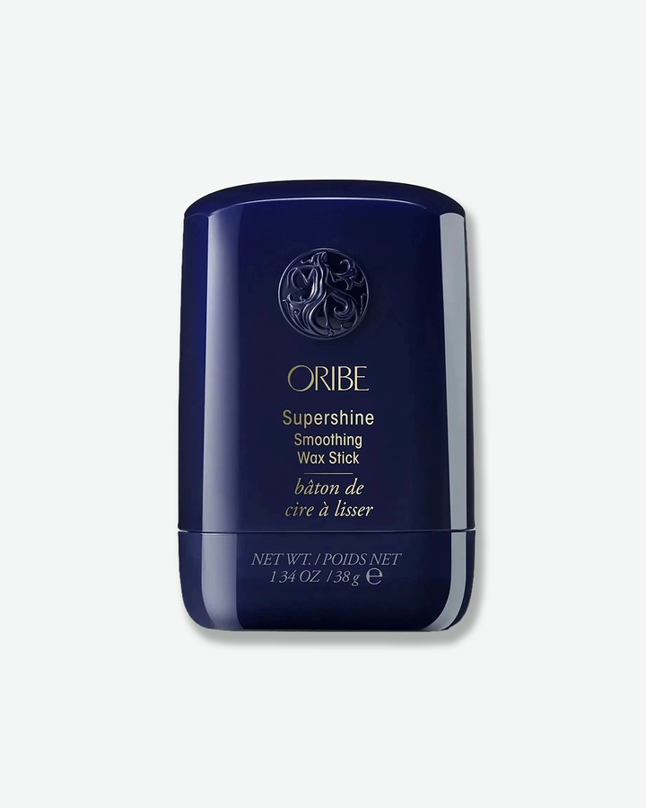 A product shot of an Oribe Supershine Smoothing Wax Stick container, centered against a plain light gray background. The curved, oval-shaped container is made of a glossy, deep midnight blue plastic with a matching pull-off cap. The upper portion of the cap features Oribe's signature circular embossed goddess emblem. Below it, the brand name "ORIBE", "Supershine Smoothing Wax Stick", and the French translation "bâton de cire à lisser" are printed in fine gold script. The smaller base twist-section at the bo