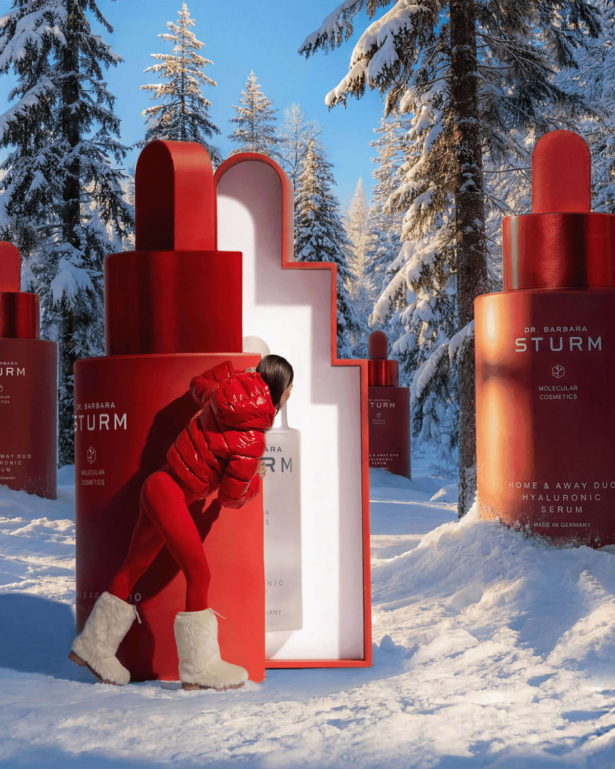 Dr. Barbara Sturm Home & Away Duo holiday campaign featuring oversized red and white Hyaluronic Serum bottles in a snowy alpine forest setting with a model dressed in red.