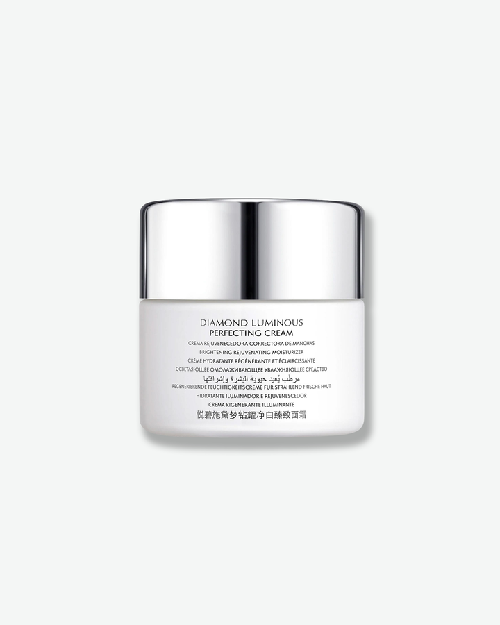 Front view of a Natura Bissé Diamond Luminous Perfecting Cream jar against a plain gray background. The matte white cylindrical jar displays detailed multi-lingual product descriptions printed in a subtle grey font across the front beneath a high-shine silver cap.
