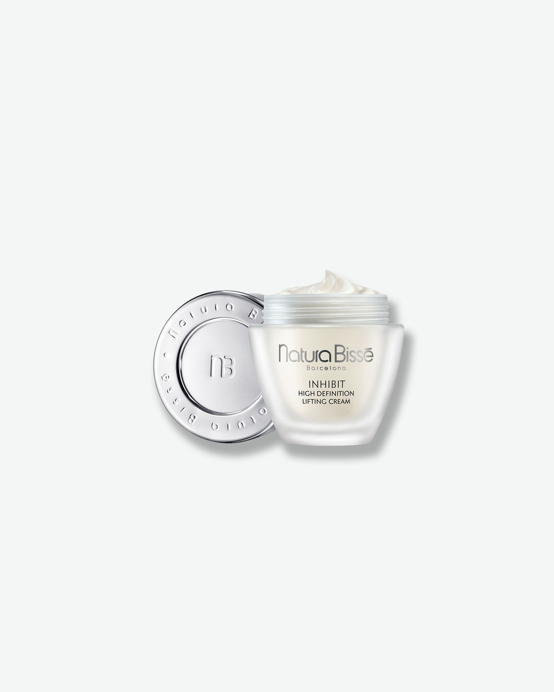 Product shot of the open Natura Bissé Inhibit High Definition Lifting Cream jar. The tapered frosted glass jar is filled with a rich white cream, showing a soft swirl on top. The detached silver cap rests behind it on its side, revealing an embossed monogram logo and the brand name around the inner lid surface.