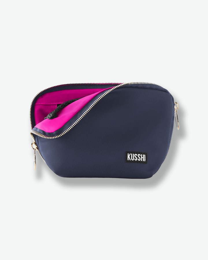 Front view of a medium navy blue fabric KUSSHI makeup bag. The gold zipper is pulled back diagonally across the front, revealing its signature hot pink interior lining and an attached black mesh detail.