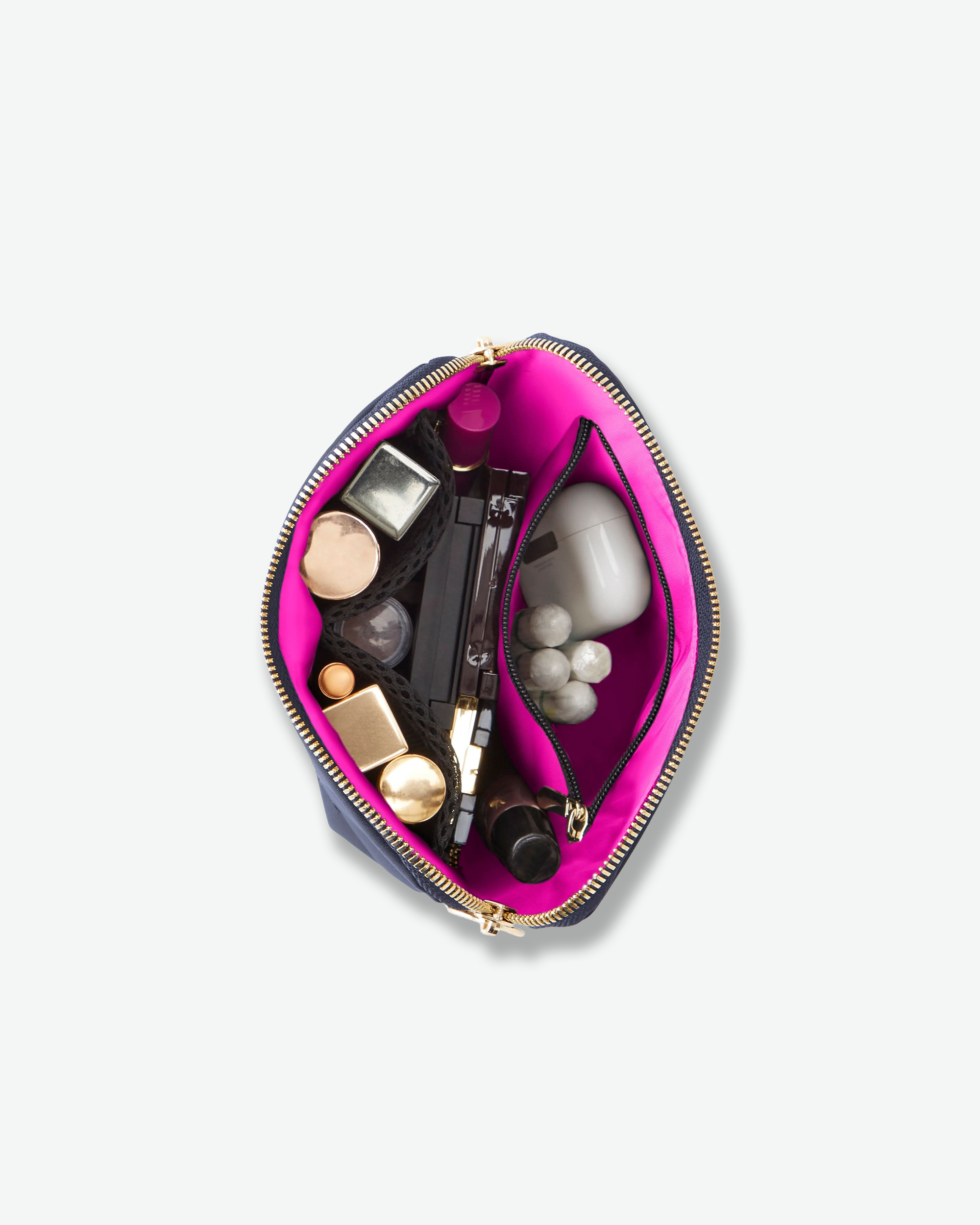 An open makeup bag with a navy blue exterior and a bright pink interior, shown from above on a light gray background. The bag is filled with various cosmetics and skincare items.