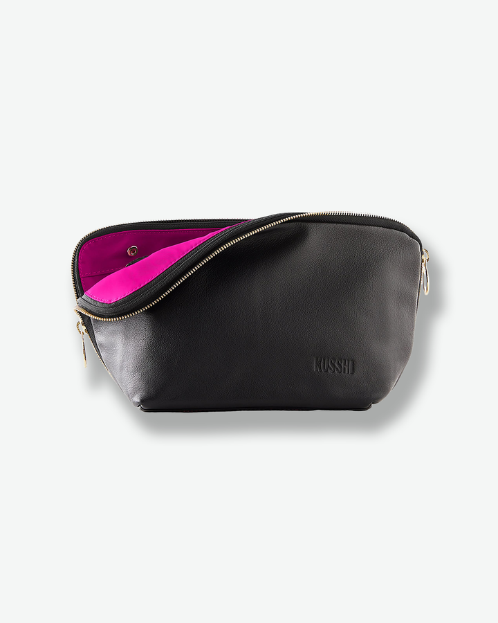 Front view of a black pebble leather KUSSHI makeup bag against a plain gray background. The gold zipper is unzipped halfway, folding down to reveal a stark contrast with its bright, hot pink fabric interior lining. The brand name is subtly debossed on the bottom right corner.