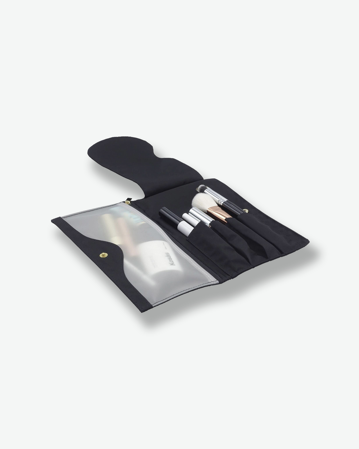 Angled view of an open black fabric makeup brush roll laid flat. The right side features specialized slots holding several cosmetic brushes, while the left side features a frosted translucent plastic zipper compartment filled with makeup products.