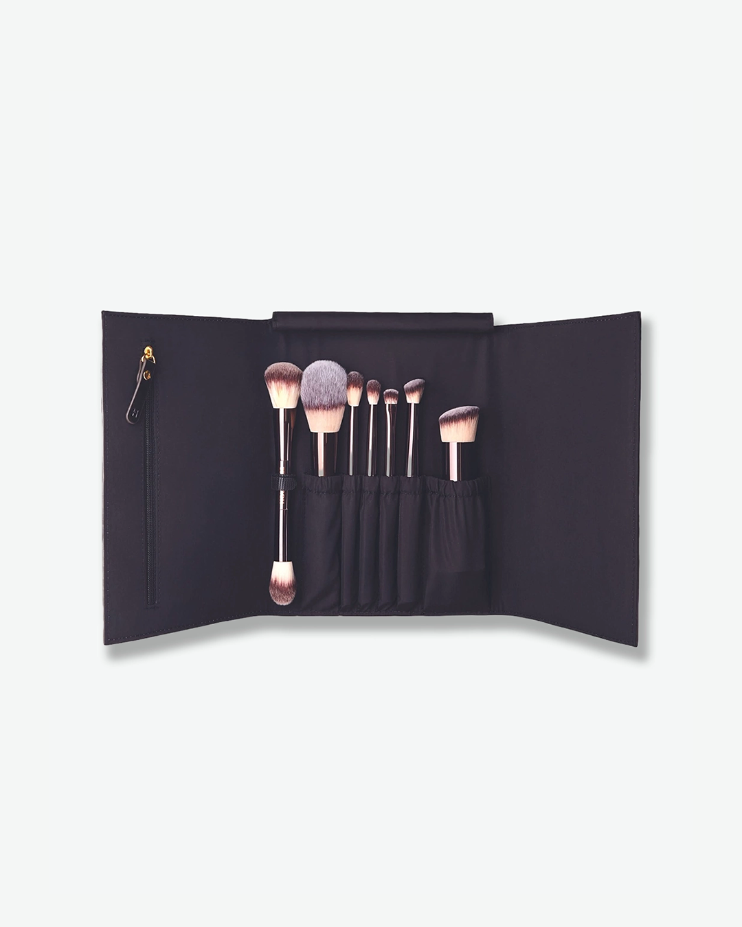 A top-down view of an open Hourglass makeup brush roll against a white background. The black fabric trifold wrap is fully extended, revealing seven essential face and eye makeup brushes with metallic handles tucked neatly into slots. A vertical zippered pocket is built into the left interior flap.