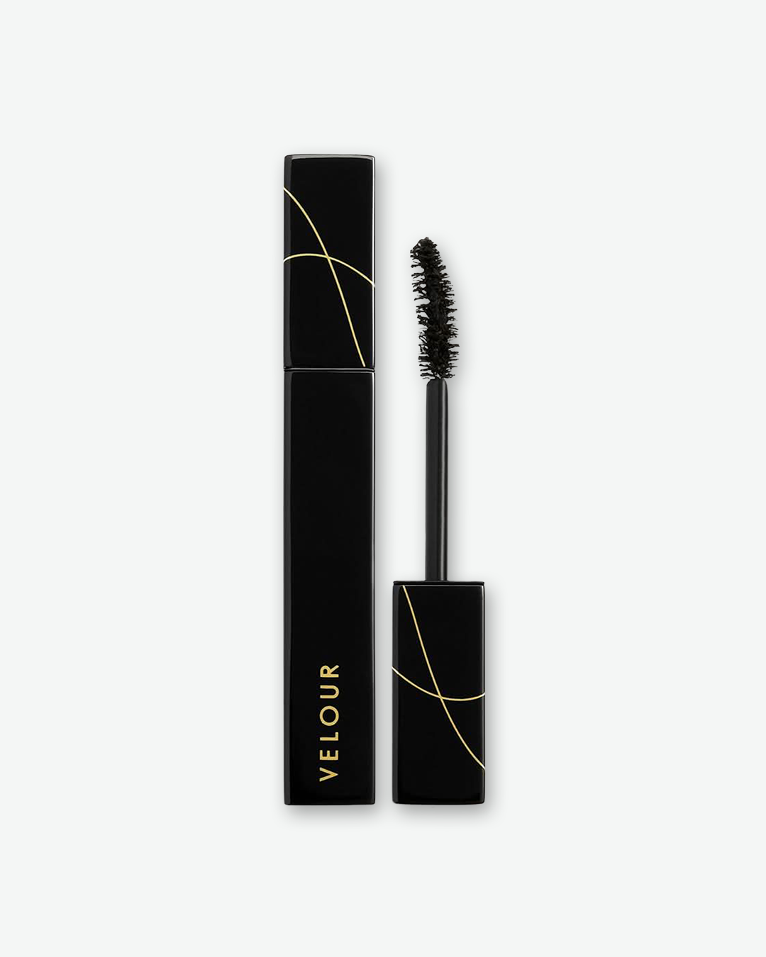 Product shot of Velour mascara against a plain gray background. On the left is the closed, square-profile black tube displaying the brand name vertically in gold. On the right, the matching wand is unscrewed, showcasing a curved black bristle brush applicator. Both sections feature abstract, thin gold line graphics crisscrossing the cap areas.