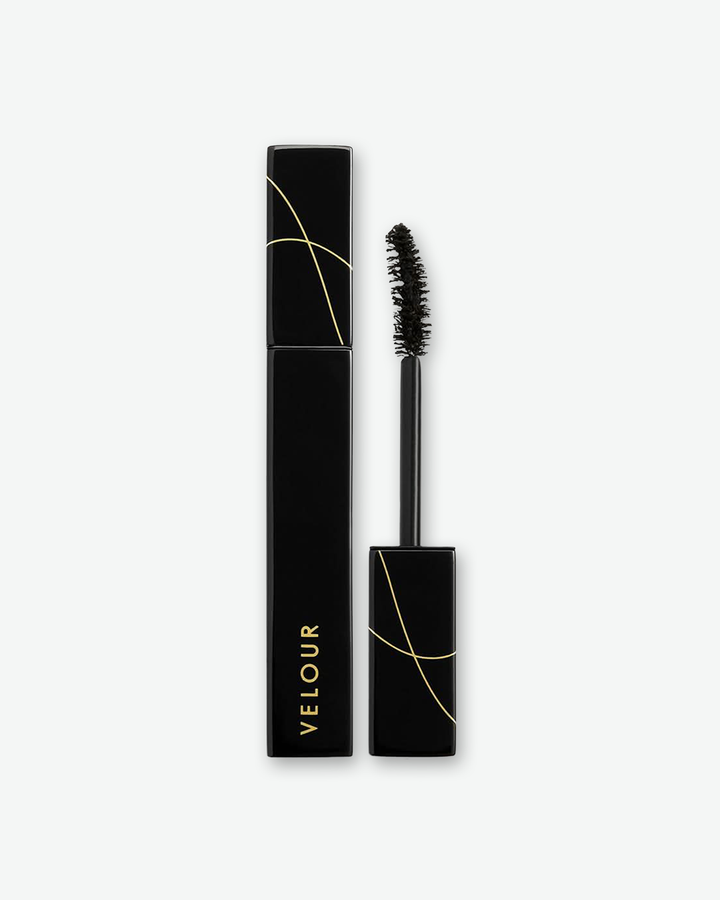 Product shot of Velour mascara against a plain gray background. On the left is the closed, square-profile black tube displaying the brand name vertically in gold. On the right, the matching wand is unscrewed, showcasing a curved black bristle brush applicator. Both sections feature abstract, thin gold line graphics crisscrossing the cap areas.