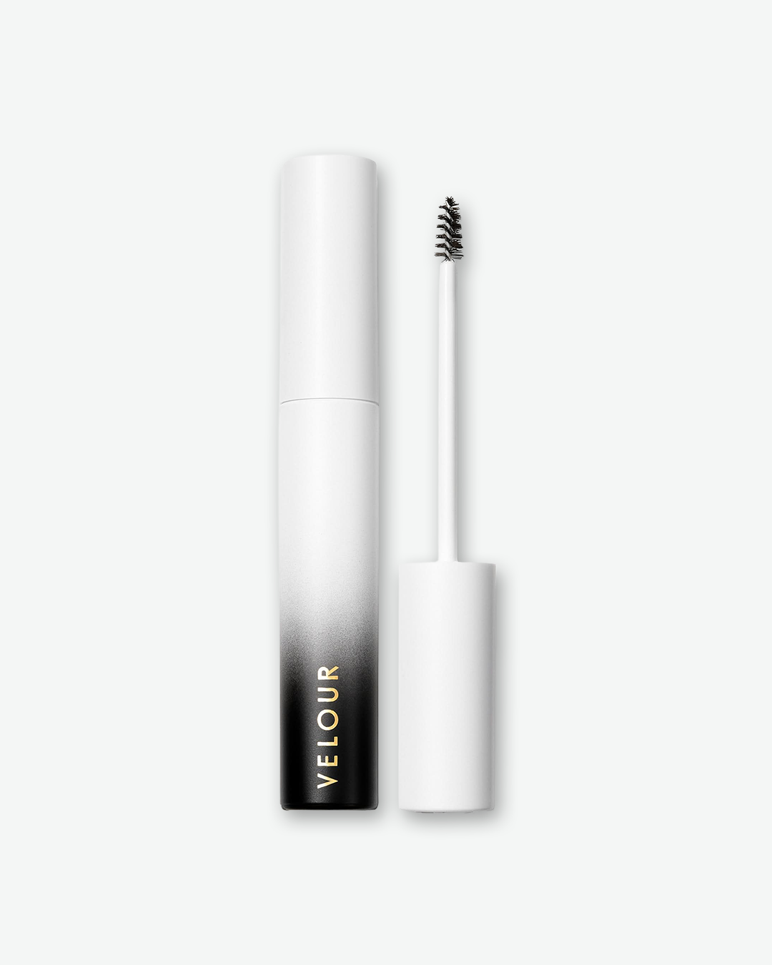 Product shot of Velour brow gel against a plain gray background. The cylindrical container features an ombre finish that fades from solid white at the top to deep black at the base, with the brand name printed vertically in gold. To the right, the unscrewed applicator wand stands vertically, revealing a small, tapered black spoolie brush.