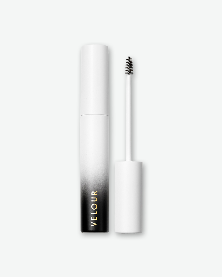 Product shot of Velour brow gel against a plain gray background. The cylindrical container features an ombre finish that fades from solid white at the top to deep black at the base, with the brand name printed vertically in gold. To the right, the unscrewed applicator wand stands vertically, revealing a small, tapered black spoolie brush.