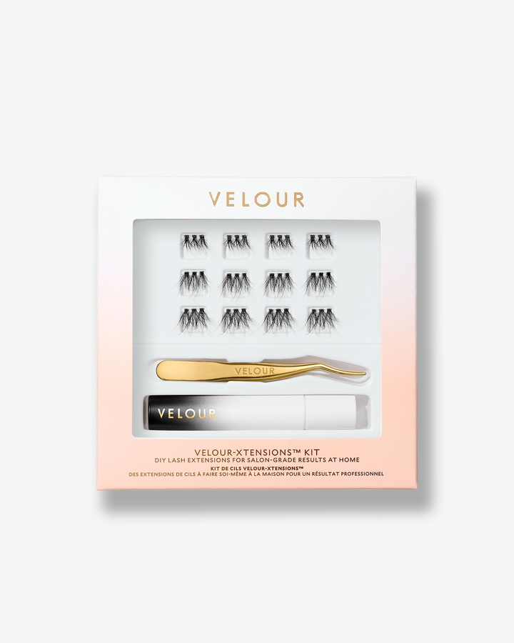 Front view of the Velour-Xtensions™ Kit against a plain gray background. The square, white-to-peach gradient gift box features a large clear window displaying a complete DIY lash extension set, containing 12 black lash clusters, a curved gold metallic applicator tool, and a small black-and-white gradient adhesive tube.