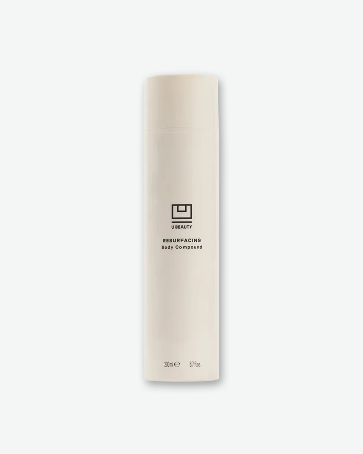 A product shot of a U Beauty Resurfacing Body Compound bottle, centered against a plain light gray background. The tall, cylindrical bottle features a soft, matte off-white finish with a matching flush cap. The black U Beauty logo emblem is printed in the center above the text "RESURFACING Body Compound", with the volume "200 mL e 6.7 fl. oz." detailed near the base.