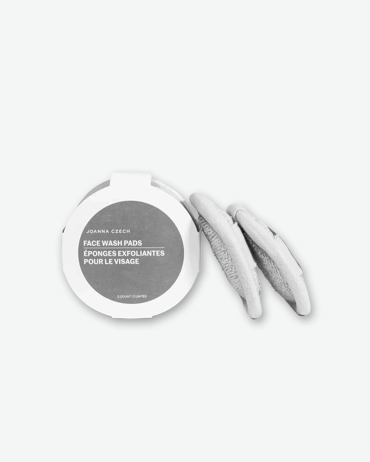 A product shot of a Joanna Czech Face Wash Pads set against a plain light gray background. On the left is the circular white cardboard sleeve packaging with a dark grey center label that reads "JOANNA CZECH", "FACE WASH PADS", and "ÉPONGES EXFOLIANTES POUR LE VISAGE". On the right, two grey terry-cloth cleansing pads are tucked slightly behind the packaging, showing their soft, textured edges.