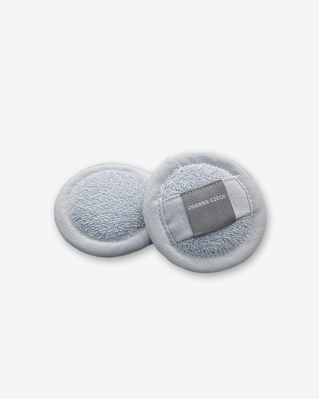 A product shot of two round Joanna Czech Face Wash Pads resting against each other on a plain light gray background. The reusable cleansing pads are made of a soft, light grey terry cloth with a fabric piping edge. The pad on the right displays its back side, featuring an integrated finger strap made of grey herringbone-patterned fabric with "JOANNA CZECH" printed in white.