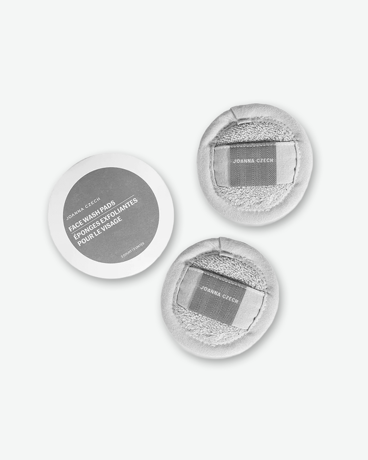 A top-down product shot displaying a Joanna Czech Face Wash Pads set, unboxed on a plain light gray background. On the left sits the round white cardboard packaging insert with its dark grey product label. To the right are two round, light grey terry-cloth cleansing pads, both flipped to show their textured finger straps detailed with grey herringbone patterns and white brand text.