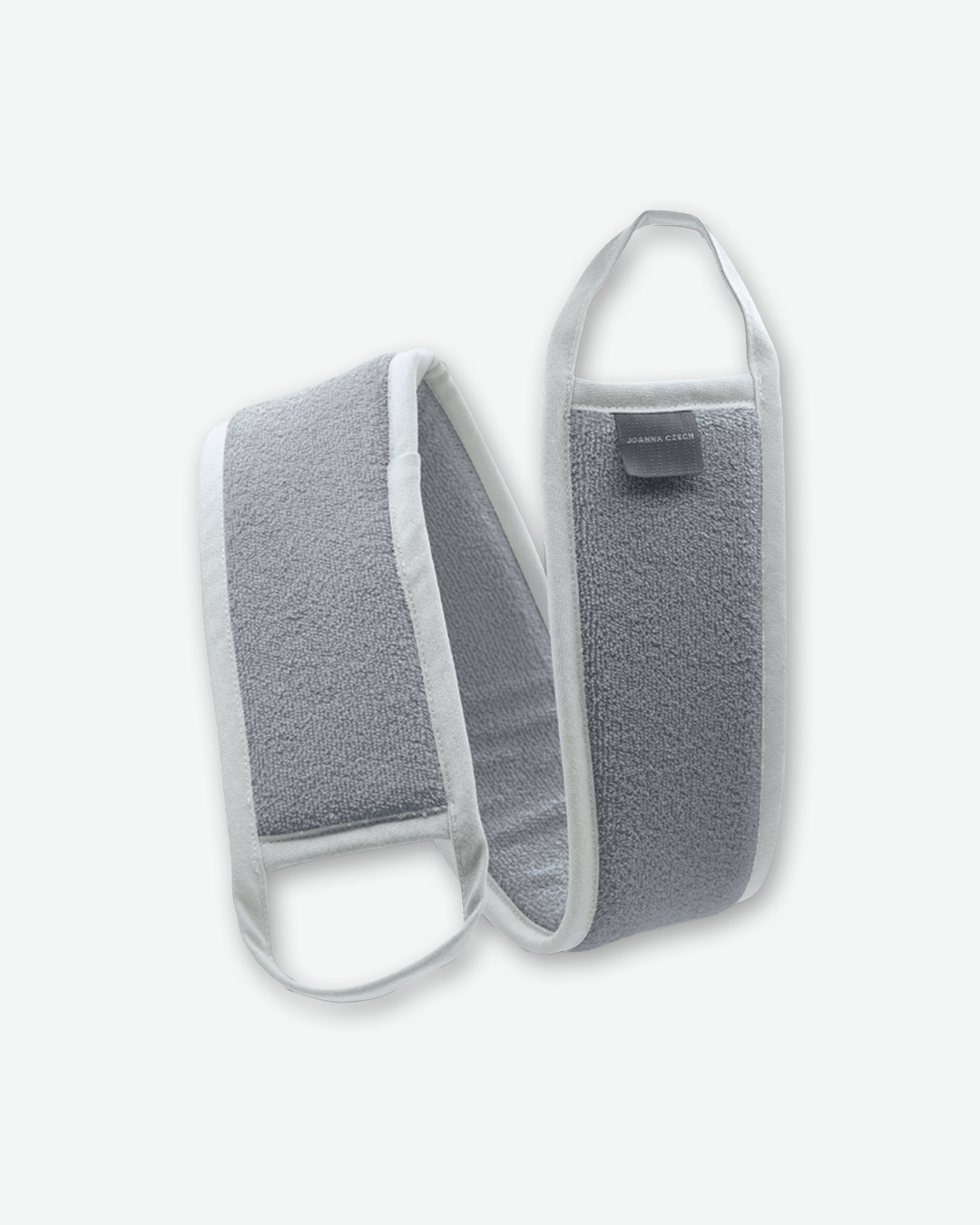 A product shot of a Joanna Czech body scrubbing towel, centered against a plain light gray background. The long, flexible strap-style towel is made of a textured, grey terry-cloth material with a clean white fabric trim bordering the edges. Each end features an integrated white fabric loop handle for gripping, and a small dark grey brand tag reading "JOANNA CZECH" is stitched onto the upper section.