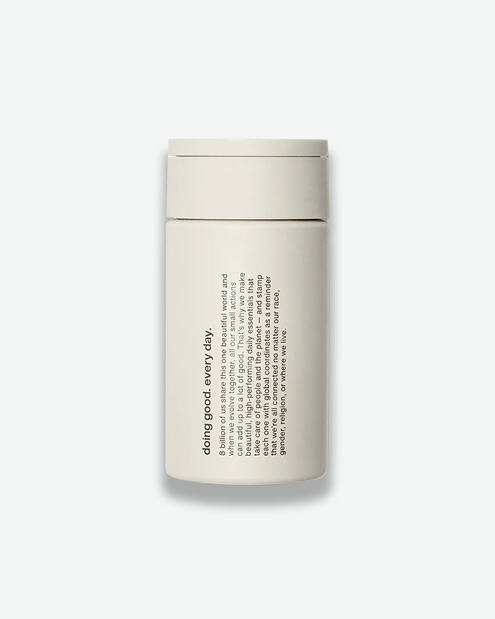A product shot of a cylindrical skincare container, centered against a plain light gray background. The bottle is a solid matte off-white color with a matching flush cap. Printed vertically on the side in clean black typography is a brand statement beginning with the bold phrase "doing good. every day." followed by a paragraph of lower-case text discussing shared global actions and coordinates.