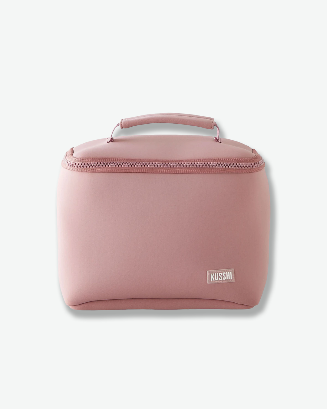 A studio front product shot of a structured, dusty rose pink fabric makeup bag from Kusshi against a light gray background. The rectangular bag features rounded sides, a matching pink zipper closure wrapping around the upper edge, and a sturdy top handle. A small, rectangular pink and white logo patch with the word 'KUSSHI' is stitched onto the bottom right corner.