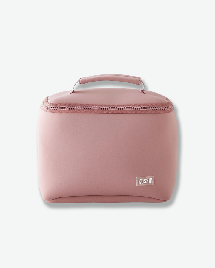 A studio front product shot of a structured, dusty rose pink fabric makeup bag from Kusshi against a light gray background. The rectangular bag features rounded sides, a matching pink zipper closure wrapping around the upper edge, and a sturdy top handle. A small, rectangular pink and white logo patch with the word 'KUSSHI' is stitched onto the bottom right corner.