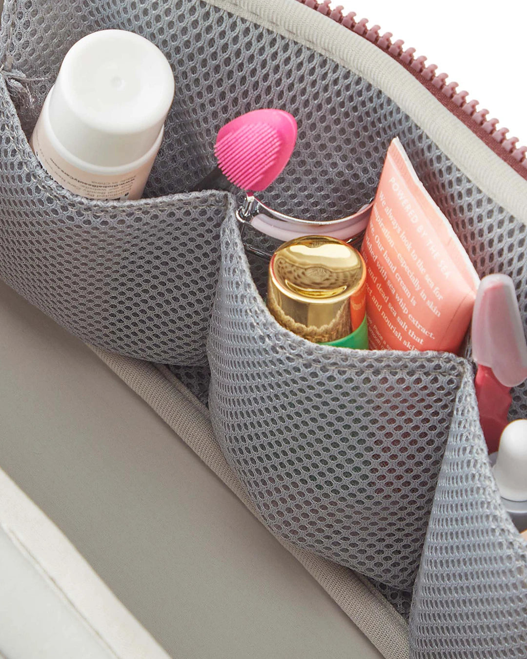 Gray mesh pouch with cosmetic items including a gold bottle and pink brush.